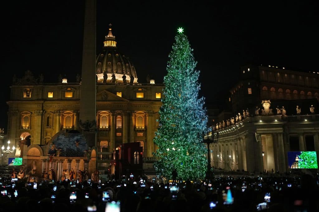 العمارة المؤقتة وشجرة عيد الميلاد الشاهقة والمضاءة بأضواء خضراء وبيضاء تقف أمام كنيسة القديس بطرس ليلاً، بينما يلتقط الحشد اللحظة بهواتفهم، مع أعمدة برنيني والمذبح الفاتيكاني الذين يحيطان بالمشهد.