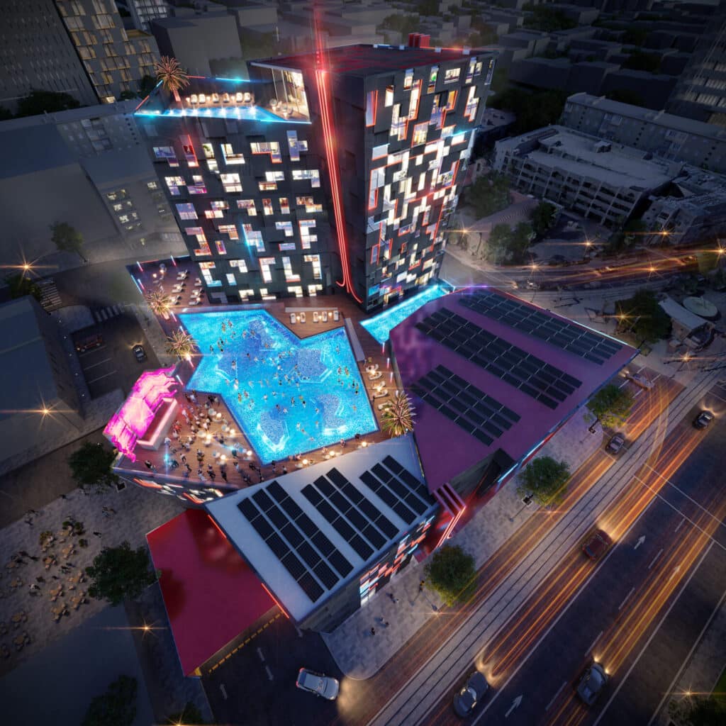 Aerial night view of the Atari Hotel in Phoenix, featuring a pixelated facade, rooftop pool shaped like a game controller, and solar panels on adjacent structures.
