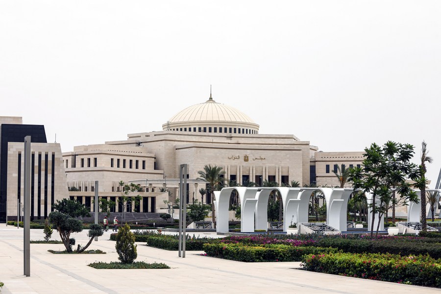 العمارة البرلمانية و منظر واسع لمبنى  البرلمان المصري الجديد في العاصمة الإدارية، محاطًا بمساحات خضراء حديثة وأقواس بيضاء تحت سماء غائمة.