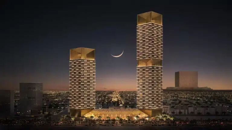 Two illuminated twin towers with golden crowns stand against a twilight sky with a crescent moon, overlooking a sprawling cityscape from their shared podium base.