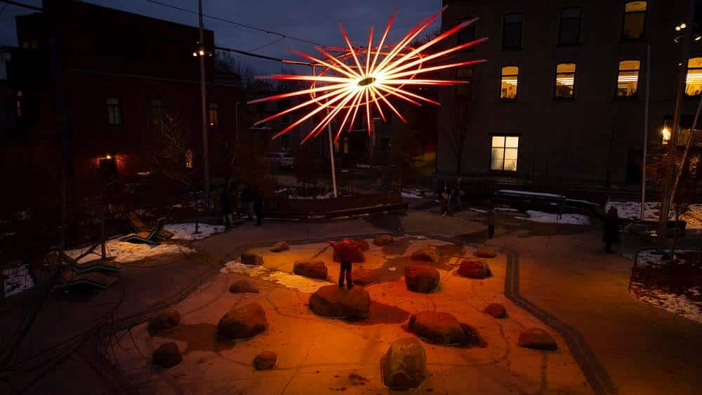 light installation glowing in red and white above Place des Fleurs-de-Macadam at night, with a child standing on rocks below and snow-dusted ground surrounding the plaza.