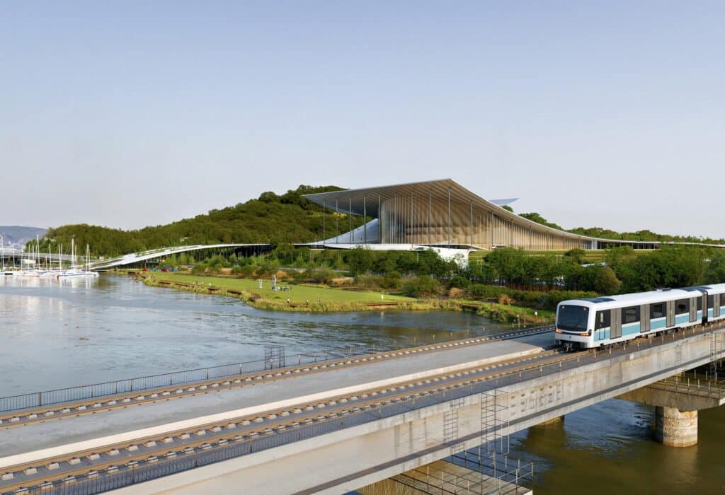 Ulsan Performing Arts Venue viewed from across the river, with a metro train crossing a bridge in the foreground. The architectural design integrates landscape, transit, and cultural infrastructure.