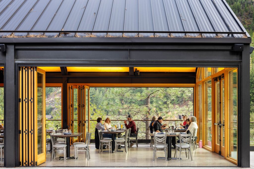 يجلس الضيوف داخل جناح ريفر إيدج مع أبواب زجاجية قابلة للطي مفتوحة على منظر غابة ونهر تحت سقف معدني داكن.
