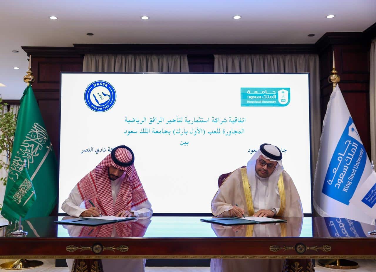 Two officials signing a memorandum of understanding for the Al-Nassr training complex at King Saud University, flanked by Saudi and university flags.