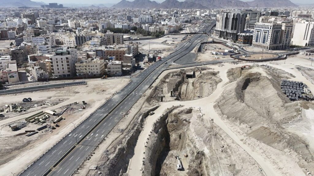 An aerial photograph showing a wide, multi-lane asphalt highway cutting through a city under development. The foreground features large excavation pits and dirt mounds with construction machinery, while the background shows a dense city layout with mountains on the horizon.