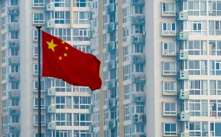 The national flag of China (a red field with five golden stars) flying in front of a tall, modern apartment building with numerous windows and balconies.