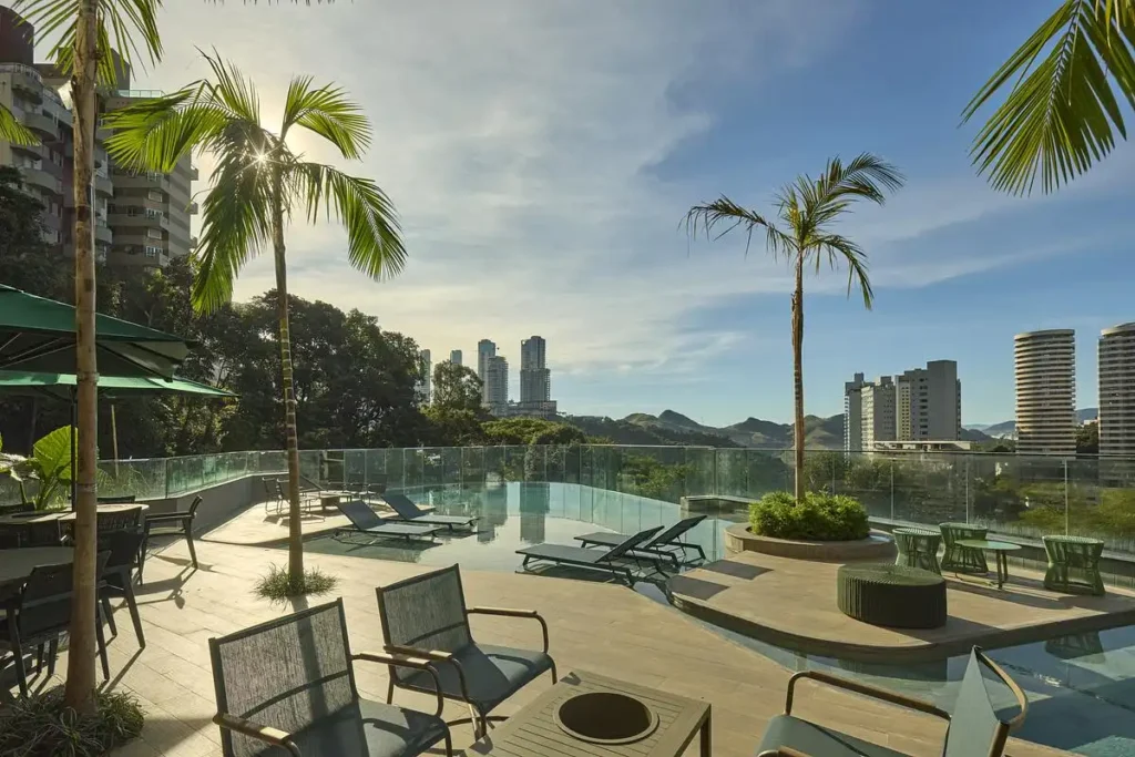 Rooftop pool terrace at Edif&iacute;cio Mir&oacute; Tower with lounge chairs, palm trees, and panoramic views of Buen&oacute;polis skyline under a bright sky.
