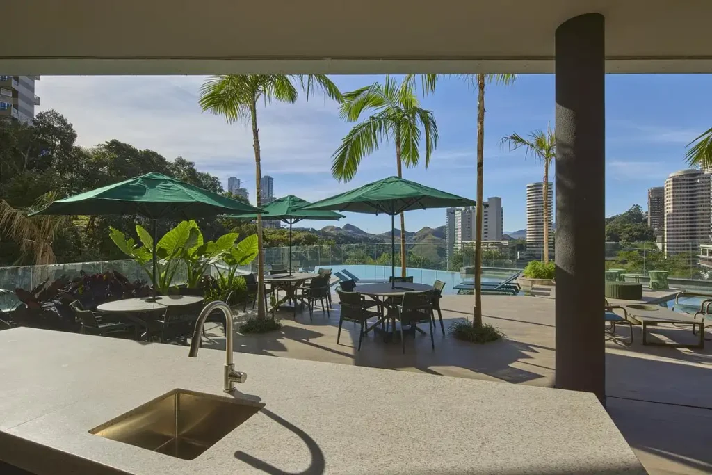 Outdoor kitchen counter overlooking the pool terrace at Edif&iacute;cio Mir&oacute; Tower, with green umbrellas, palm trees, and distant city skyline under a bright sky.