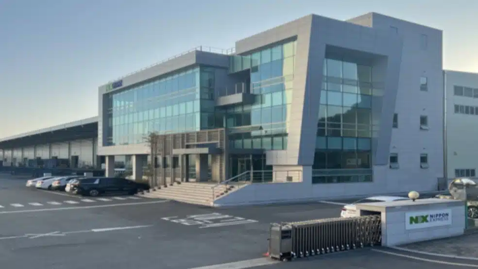 A modern multi-story glass office building and large warehouse facility for Nippon Express (NX) with a parking lot and entrance gate.