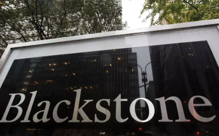 A close-up, low-angle shot of a sleek black and silver sign featuring the "Blackstone" company logo, reflecting city buildings and trees on its surface