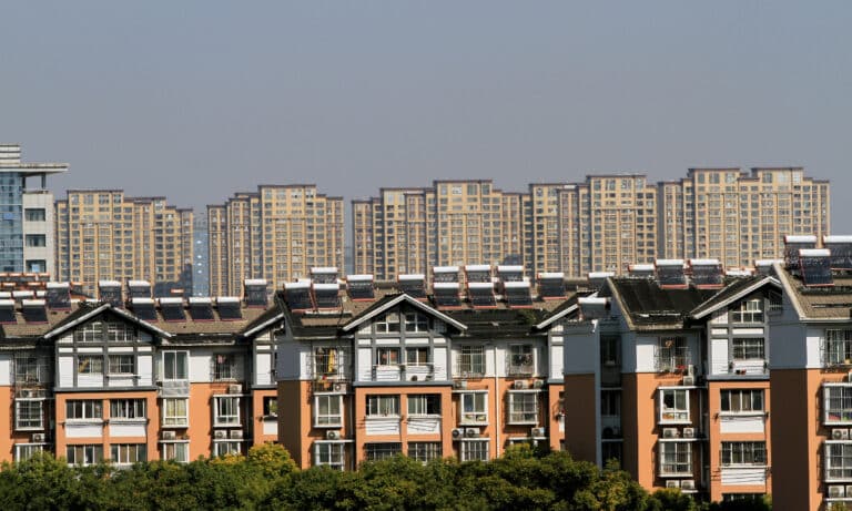 A high-angle view of multi-story residential buildings with orange and white facades, featuring numerous solar water heaters installed on their sloped roofs, with a background of dense high-rise apartment complexes under a clear sky.