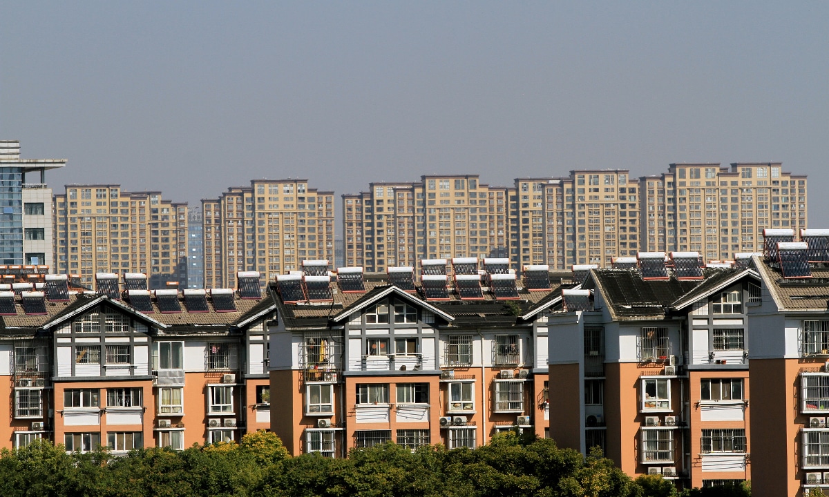 A high-angle view of multi-story residential buildings with orange and white facades, featuring numerous solar water heaters installed on their sloped roofs, with a background of dense high-rise apartment complexes under a clear sky.