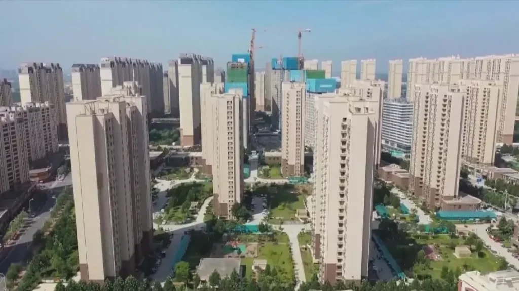 An aerial drone view of a massive residential complex featuring numerous beige high-rise apartment buildings with green spaces, walking paths, and construction cranes in the background.