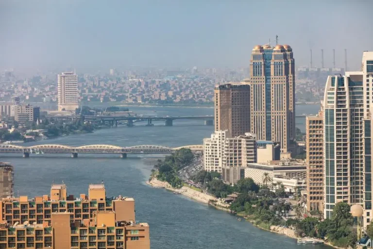 High-angle panoramic view of the Nile River in Cairo, Egypt, featuring the iconic Nile City Towers, bridges, and a dense cityscape under a clear sky.