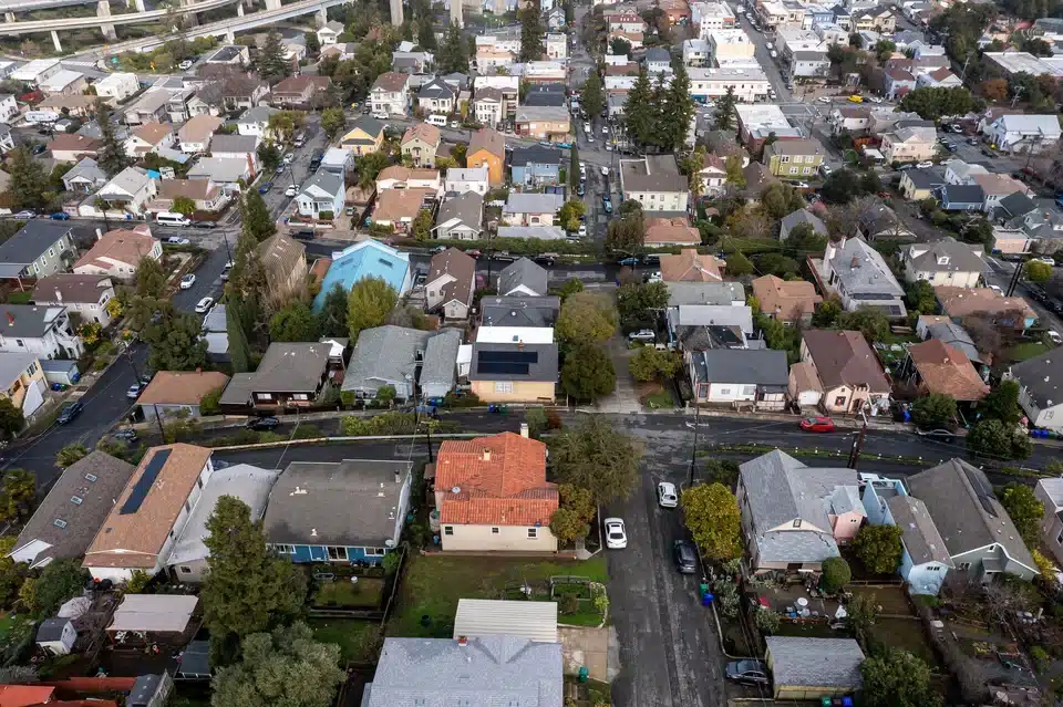An aerial, top-down view of a densely packed residential neighborhood with various colored houses, paved streets, parked cars, and scattered trees.