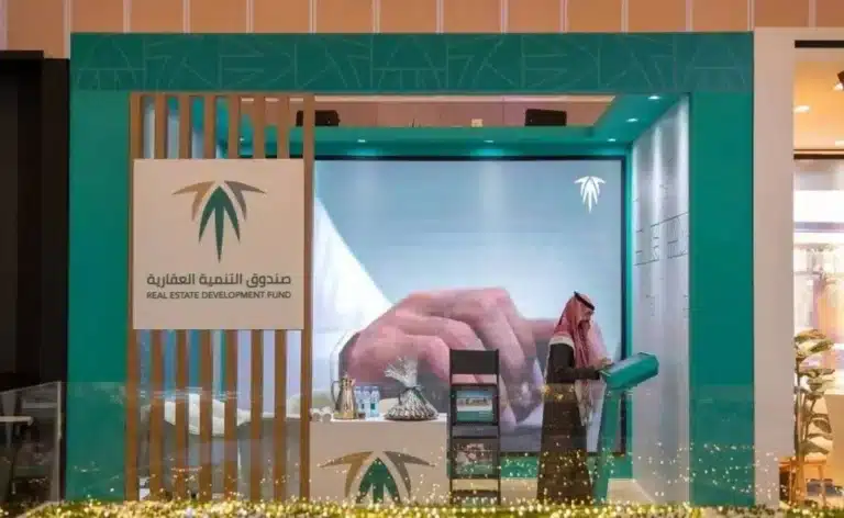 An exhibition booth for the Saudi Real Estate Development Fund (REDF) in teal and white, featuring a digital kiosk and a staff member in traditional Saudi attire.