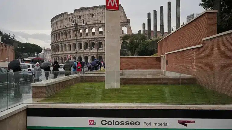 A view of the ancient Roman Colosseum in the background with a modern Metro station entrance sign for 'Colosseo Fori Imperiali' in the foreground on a rainy day with people carrying umbrellas.