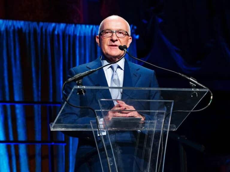 A medium shot of Albert Behler, a man with glasses and a blue suit, speaking at a transparent podium during an event with blue curtains in the background.