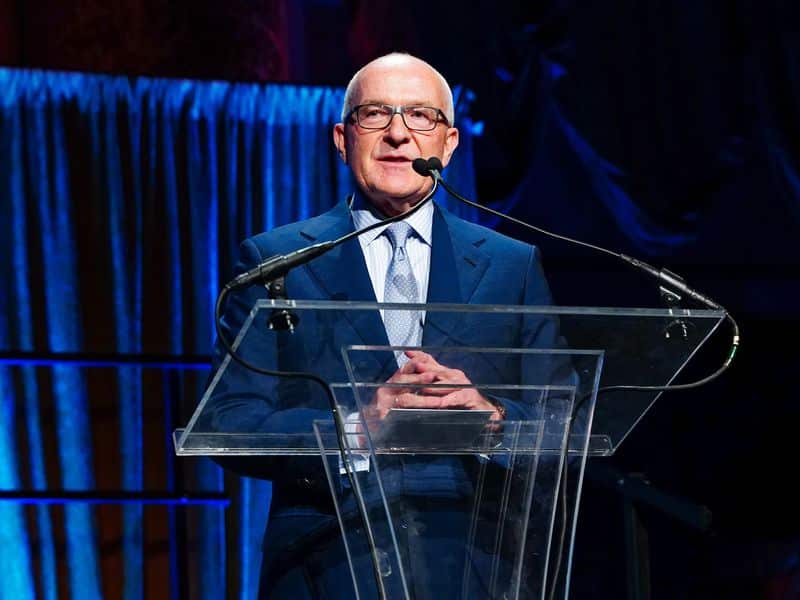 A medium shot of Albert Behler, a man with glasses and a blue suit, speaking at a transparent podium during an event with blue curtains in the background.