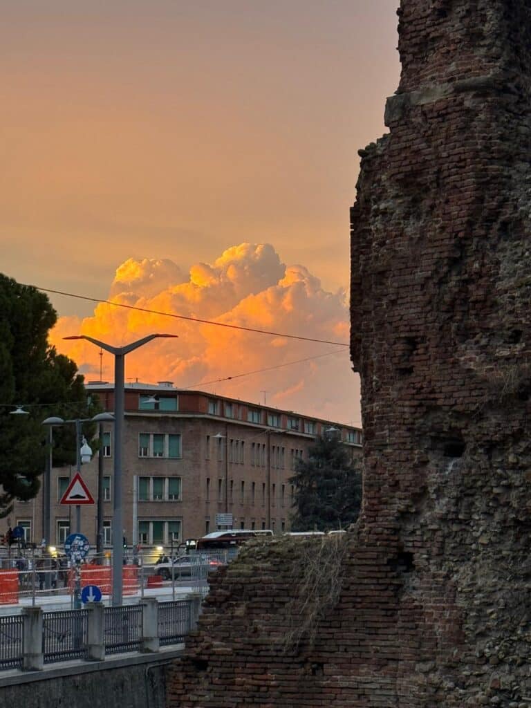 Ancient red brick ruin wall foregrounding modern residential buildings under dramatic golden sunset clouds.