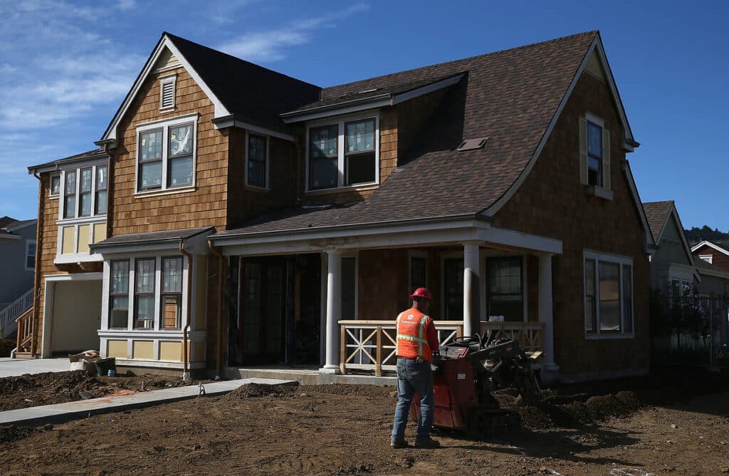 A construction worker in a red hard hat and orange safety vest operates machinery in the dirt lot in front of a newly built, two-story house with brown shingle siding and a dark brown roof.