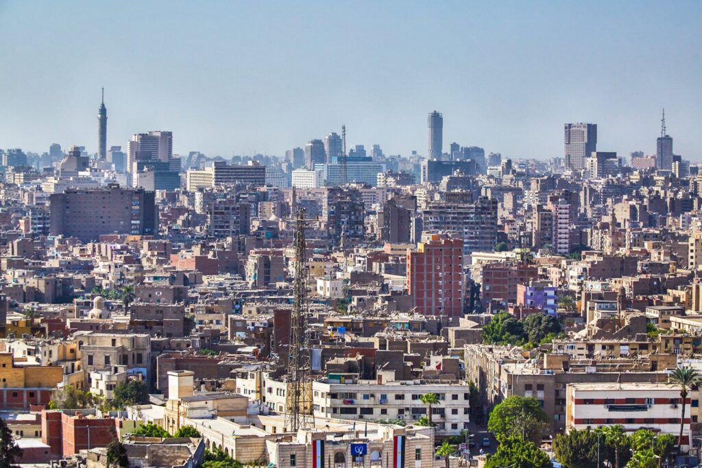 A high-angle, wide-view photograph of Cairo's dense urban landscape, showing a sea of tan and brown residential buildings with the Cairo Tower and modern skyscrapers visible on the distant horizon under a clear blue sky.