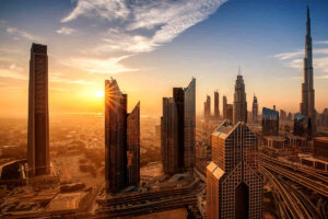 An aerial view of Dubai's skyline at sunset, showing the Burj Khalifa and modern skyscrapers with a bright sunburst reflecting off a glass tower.