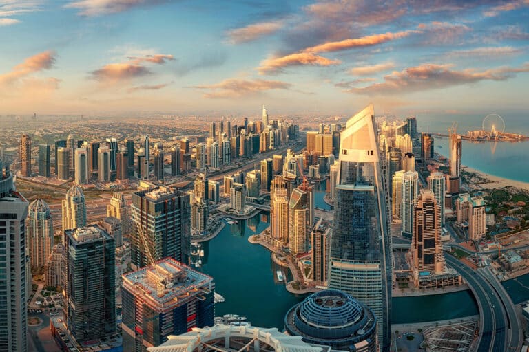 A vibrant aerial panorama of Dubai Marina at sunset, featuring glass skyscrapers, a blue winding canal with yachts, and the Ain Dubai observation wheel in the distance against a pink-tinted cloudy sky.