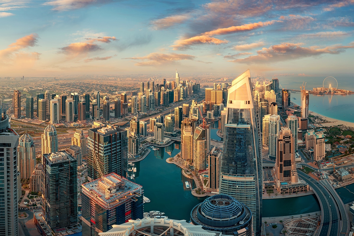 A vibrant aerial panorama of Dubai Marina at sunset, featuring glass skyscrapers, a blue winding canal with yachts, and the Ain Dubai observation wheel in the distance against a pink-tinted cloudy sky.