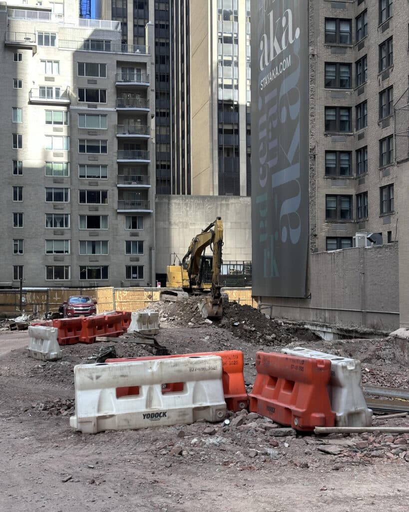 A yellow excavator sits on a dirt mound at a construction site surrounded by tall apartment buildings and orange and white plastic barriers.