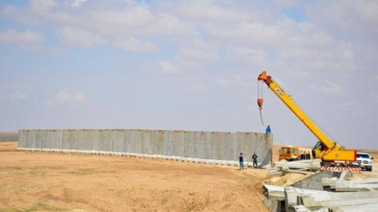 construction site in a desert landscape showing a long row of concrete T-wall blast barriers being installed by a yellow mobile crane under a blue sky.
