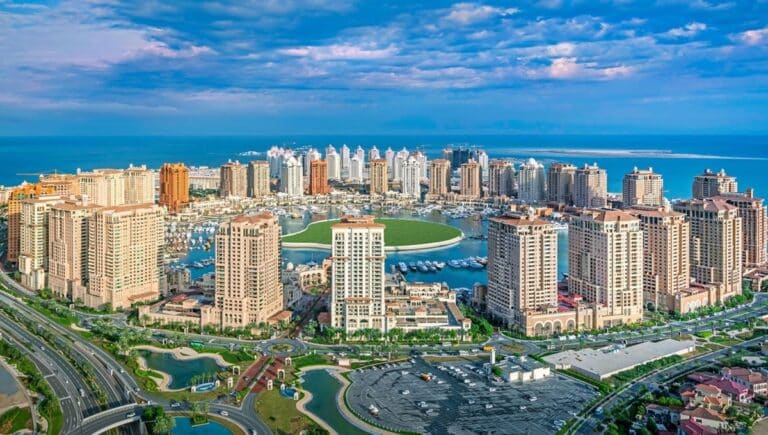 An aerial panorama of Porto Arabia at The Pearl-Qatar, featuring a circular marina filled with yachts, surrounded by Mediterranean-style high-rise luxury apartments and the turquoise sea under a blue sky.