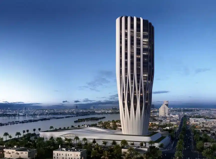 A futuristic, high-rise skyscraper with a tapering base and a white exoskeleton lattice design, situated on the banks of the Tigris River in Baghdad at dusk.