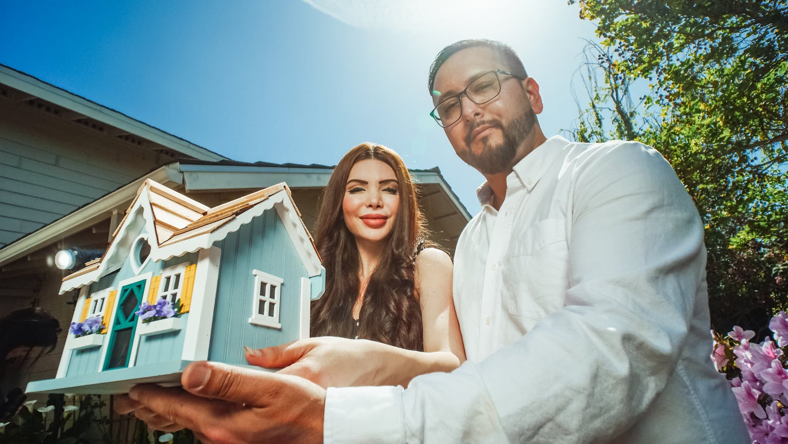A happy couple holding a miniature house model outdoors, symbolizing new home ownership.