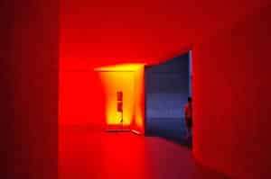 A minimalist room bathed in red light with a silhouette standing in the doorway.