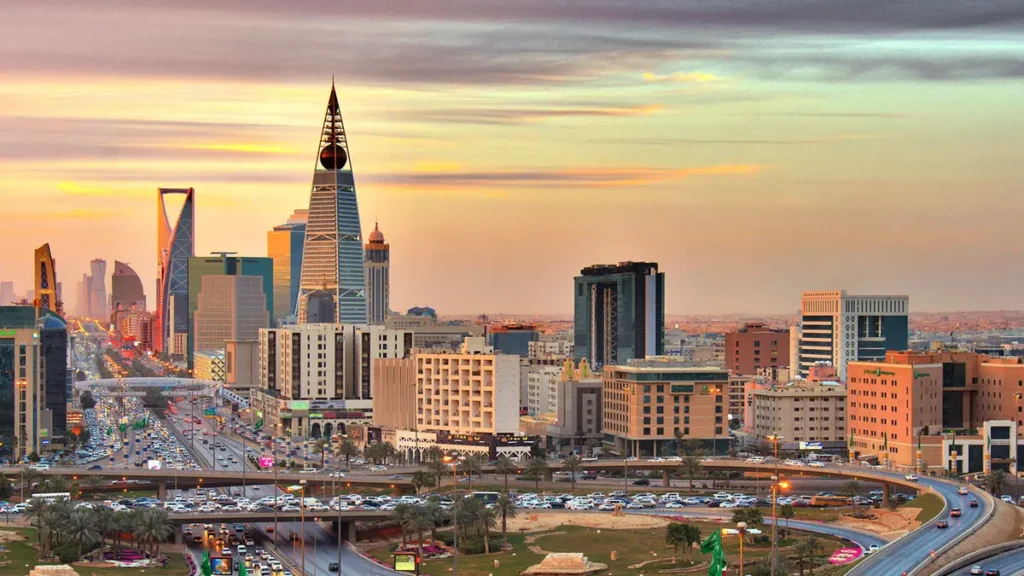 A panoramic view of Riyadh's skyline at sunset, featuring the Kingdom Centre skyscraper (spire) and the Faisaliah Tower (pyramid shape) above a busy highway (King Fahd Road).