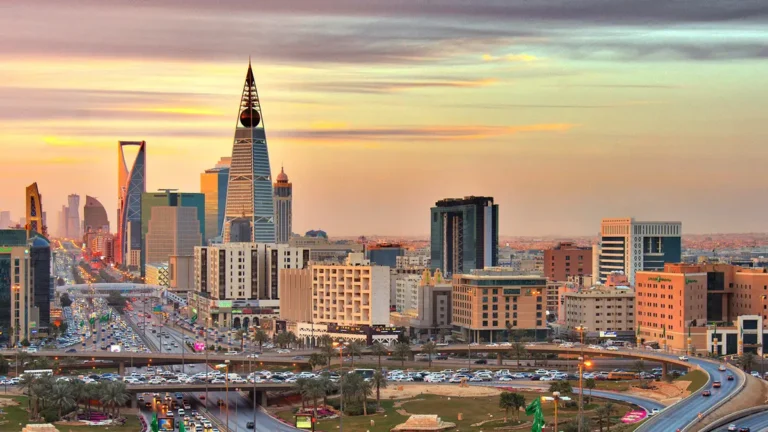 A panoramic view of Riyadh's skyline at sunset, featuring the Kingdom Centre skyscraper (spire) and the Faisaliah Tower (pyramid shape) above a busy highway (King Fahd Road).