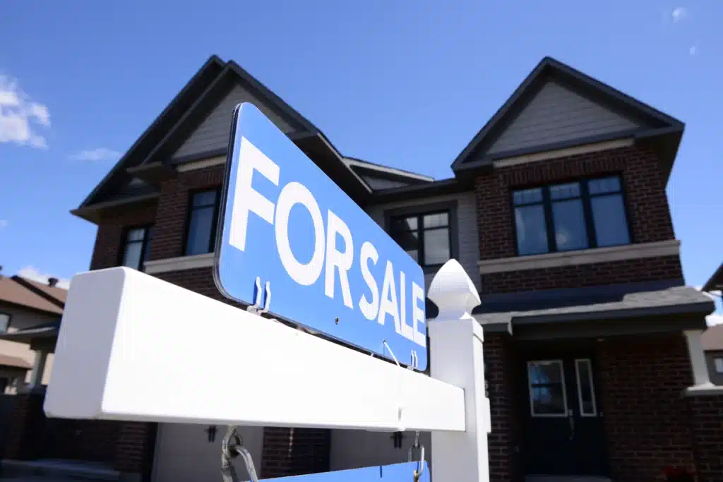 Close-up of a blue "For Sale" sign in front of a modern two-story brick residential house under a clear blue sky.