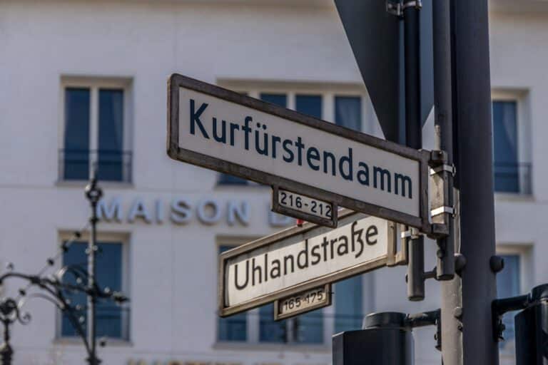 A close-up shot of two traditional Berlin street signs on a metal pole, indicating "Kurfürstendamm" and "Uhlandstraße" with building numbers below each.