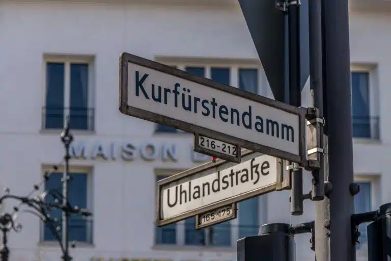 A close-up shot of two traditional Berlin street signs on a metal pole, indicating "Kurfürstendamm" and "Uhlandstraße" with building numbers below each.