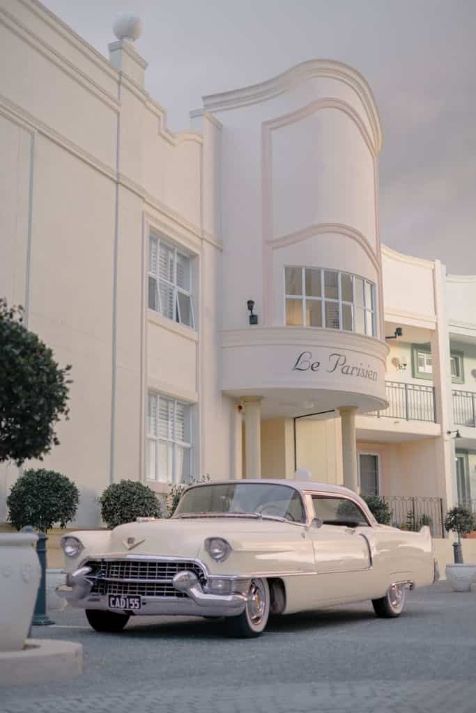 Classic white Cadillac parked outside an Art Deco building with vintage style and overcast sky.