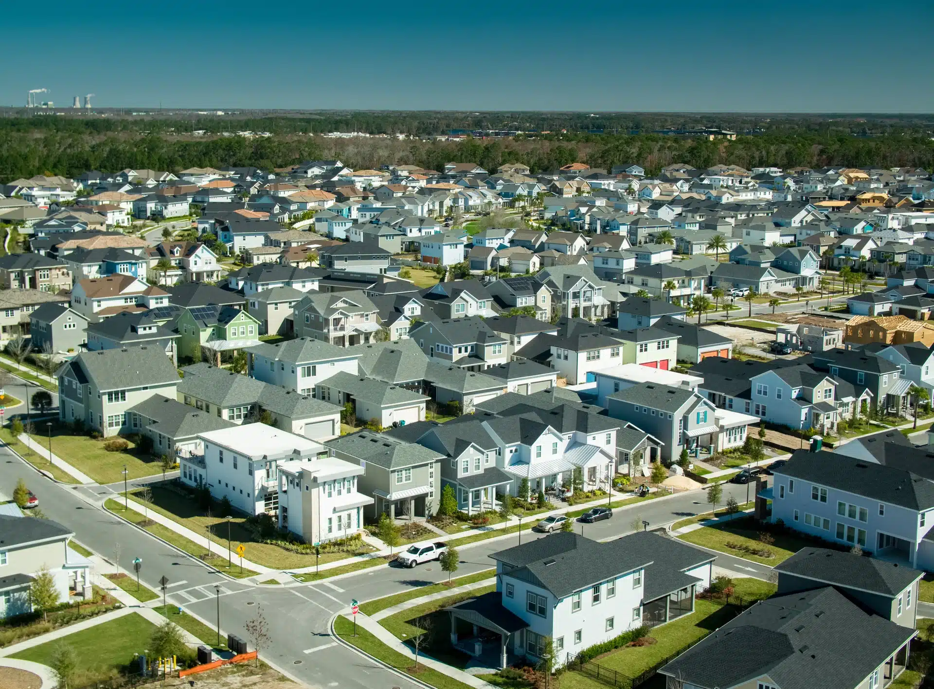 An aerial view of a dense, newly built suburban housing development featuring rows of identical and similar single-family homes with varying roof colors and façades, bordered by a large expanse of forest.