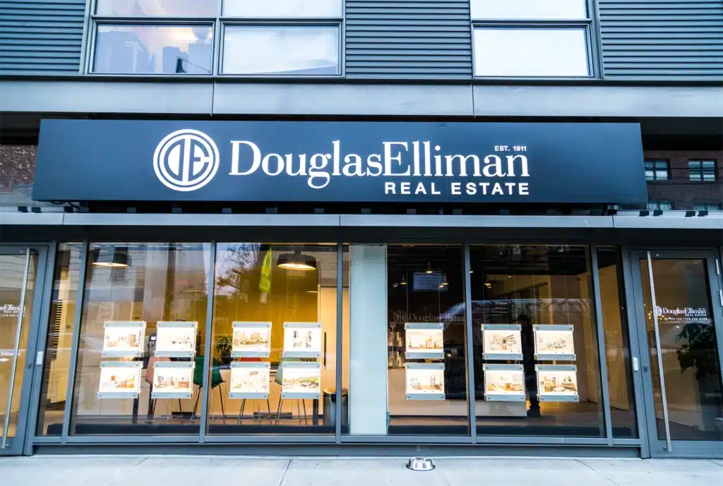 Exterior view of a Douglas Elliman Real Estate office storefront with a large black sign, featuring property listings displayed in the glass windows.