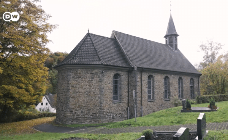 Exterior view of a historic rural stone church featuring rough masonry walls, a steep slate roof, and a small bell tower, surrounded by a peaceful landscape.