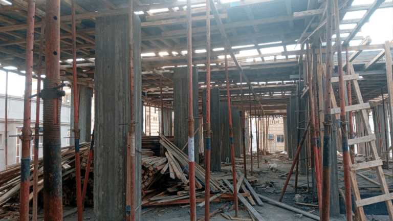 Interior view of an active construction site showing reinforced concrete columns, wooden slab formwork, and steel shoring props.