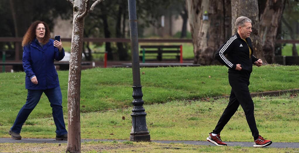 A candid, outdoor photo of a woman holding a phone walking behind a man who is jogging on a paved path in a park