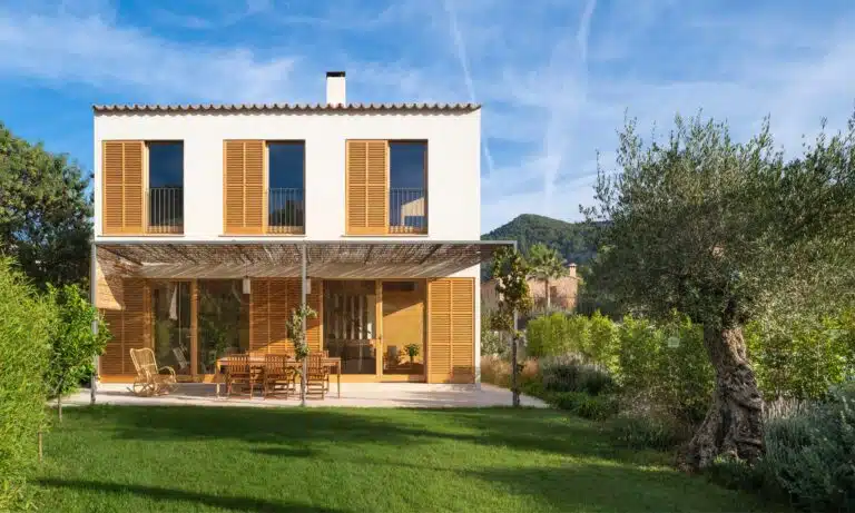 Rear facade of Puigbonnyent House showing passive solar design with wooden shutters and a shaded terrace overlooking a sustainable Mediterranean garden.