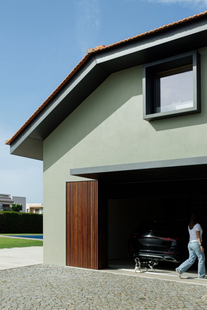 Architectural detail of Casa do Engenho showing a protruding black window frame on a sage green wall and a wooden garage door.