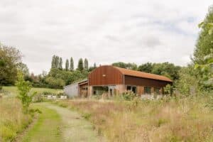 Side view of the Dutch Barn house in a wild meadow under a cloudy sky.