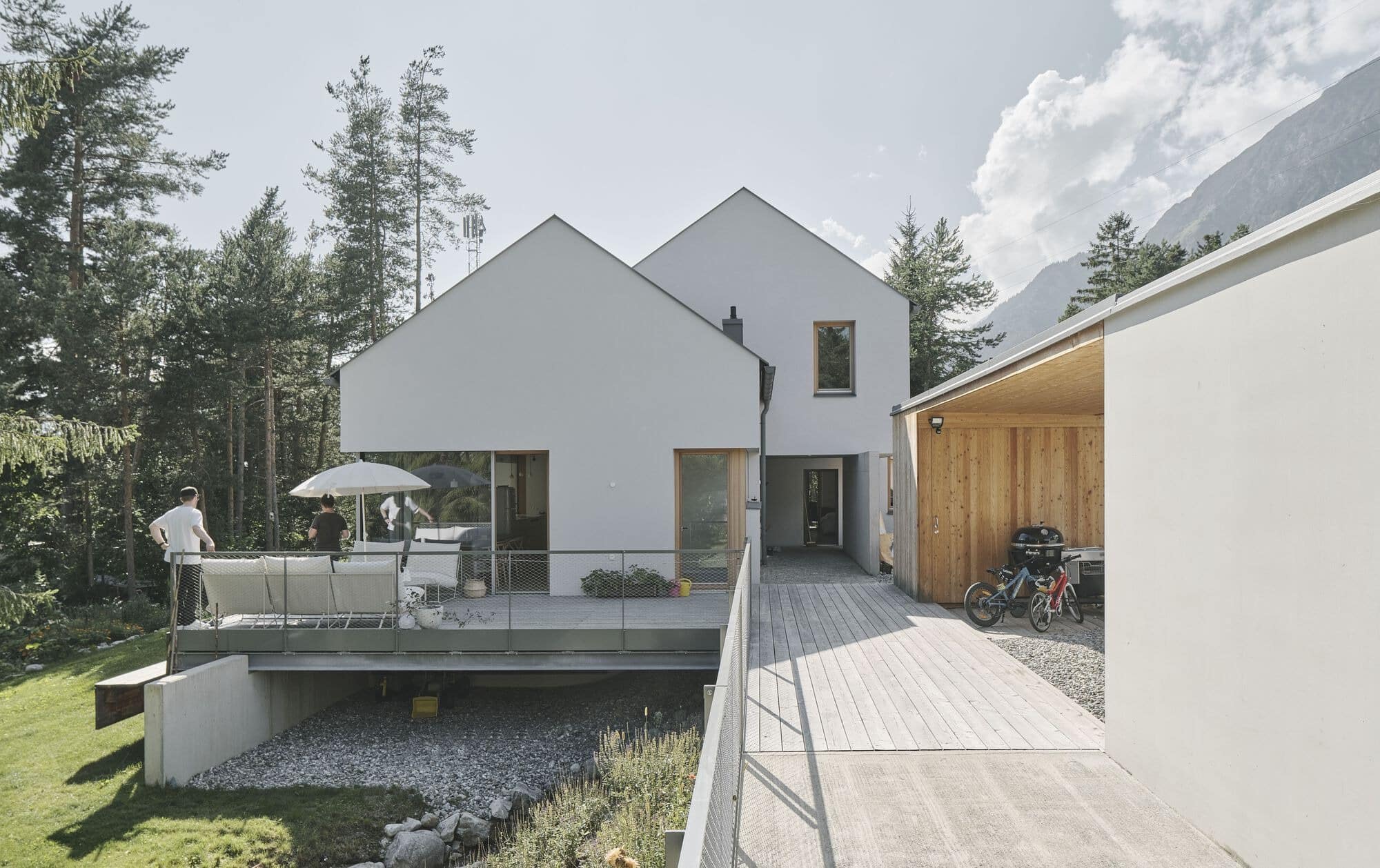 Exterior view of a modern house with white gabled roofs and a wooden deck overlooking a garden and mountains.